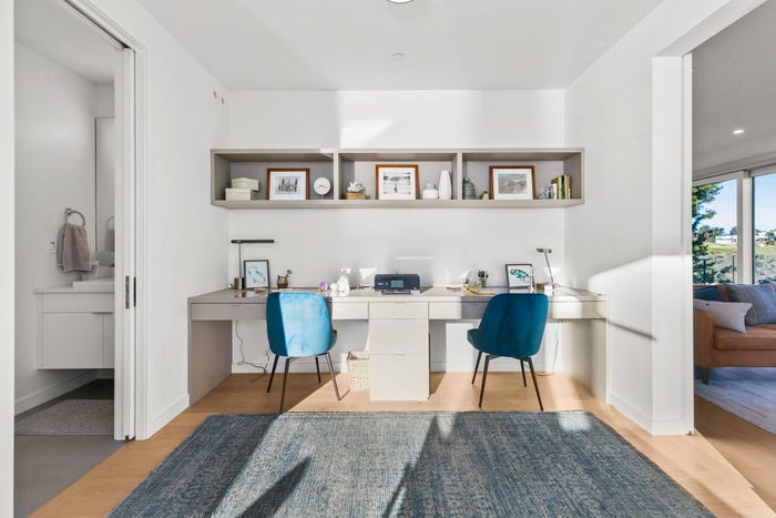 prefab home study with white two-person desk and two blue mod chairs, a shelf above with picture frames and a gray rug, dvele malibu